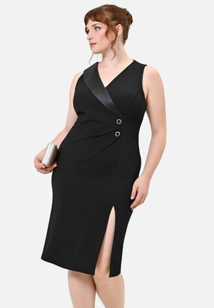 Donna con capelli rossi raccolti in uno chignon indossa un abito nero senza maniche con colletto in raso, bottoni laterali, spacco sulla coscia e tiene una pochette argentata.