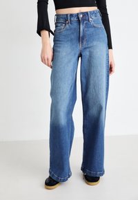 High-waisted, wide-leg jeans made of medium-wash denim. Features five pockets, copper rivets, and a subtle fade along the legs.