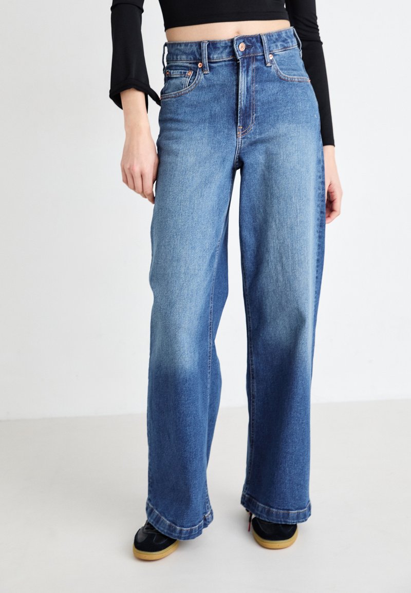 High-waisted, wide-leg jeans made of medium-wash denim. Features five pockets, copper rivets, and a subtle fade along the legs.