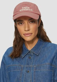 Casquette en coton rose clair avec "bonjour week-end" brodé en blanc. Portée avec une veste en denim bleu, dotée de boutons-pression et de détails de couture.