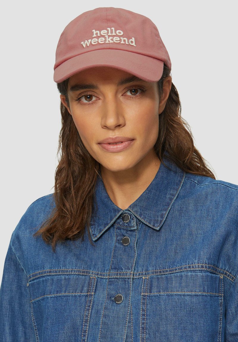 Casquette en coton rose clair avec "bonjour week-end" brodé en blanc. Portée avec une veste en denim bleu, dotée de boutons-pression et de détails de couture.