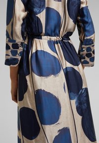 Long-sleeve dress with a cinched waist, features large navy blue dots on a beige background, smooth fabric, and button accents on sleeves.
