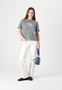 Grå bomulls t-shirt med en logotyp, matchad med vita utsvängda jeans. Hon håller en liten blå handväska och bär svarta skor. Minimalistisk bakgrund.