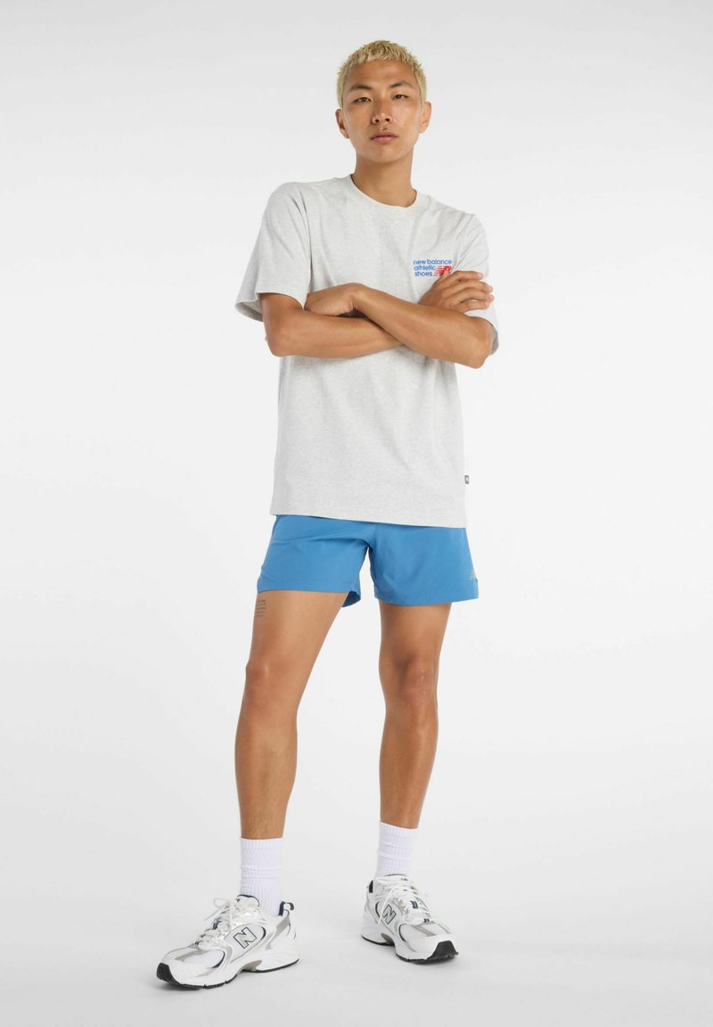 Ung man med kort blond hår står med armarna i kors iförd grå New Balance T-shirt, blå sportshorts, vita strumpor och vita sneakers.