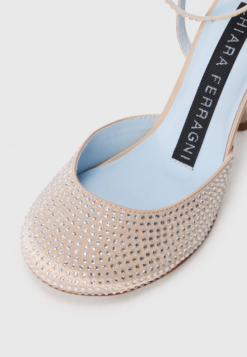 Chaussure à talons beige avec des brides autour des chevilles, recouverte de petites rivets en argent, et semelle intérieure bleu clair avec une étiquette de marque noire.