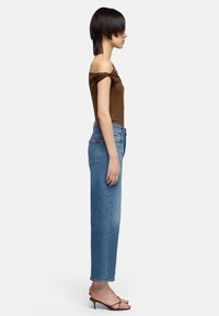 Personne de profil portant un haut marron à épaules dénudées, un jean bleu taille haute et des talons marron à lanières sur un fond blanc.