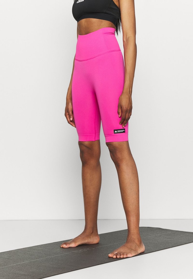 Femme debout pieds nus sur un tapis de yoga portant un short cycliste taille haute rose vif et un soutien-gorge de sport noir.