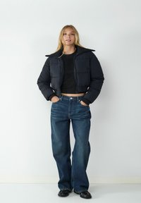 Czarna krótka kurtka puchowa z wysokim kołnierzem, zestawiona z niebieskimi dżinsami o szerokich nogawkach i wysokim stanie. Gładka faktura; minimalny design.