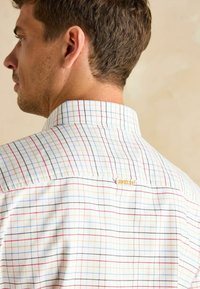 Košeľa s krátkym rukávom s kockovaným vzorom na bielom pozadí, s modrými, červenými a žltými pruhmi. Malé zlaté logo prešité na golieri.