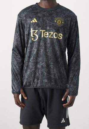 Personne portant un maillot à manches longues de Manchester United noir et gris avec des logos dorés, ainsi qu'un short Adidas noir à rayures blanches.