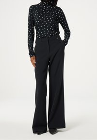 Kvinna som bär svarta vida byxor, svart polotröja med vitt stjärnmönster och svarta spetsiga skor med silvernitar, står med en hand i fickan.