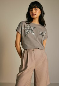 T-shirt en coton gris avec des motifs d'étoiles en sequins, manches courtes, associé à un pantalon taille haute beige clair avec des plis et des poches latérales.