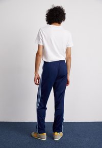 Person med lockigt hår som bär en vit t-shirt, marinblå träningsbyxor med ljusblå ränder och beige sneakers, vänd bort från kameran på en blå matta.