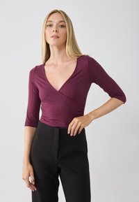 Burgundy wrap bodysuit with a V-neck, 3/4 sleeves, and soft, stretchy fabric. Paired with high-waisted black pants.