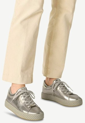Osoba nosící kovové stříbrné tenisky s texturovanými béžovými podrážkami a světlé béžové kalhoty, stojící na neutrálním pozadí.