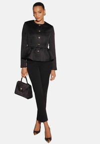 Černá peplum bunda se zlatými knoflíky, zkombinovaná s černými úzkými kalhotami a černou kabelkou s jemnou texturou a zlatými detaily.