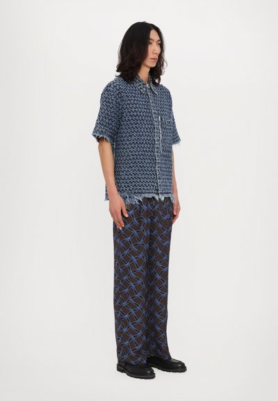 Blue woven short-sleeve shirt with a pocket, paired with brown patterned pants featuring light blue geometric lines, black footwear.