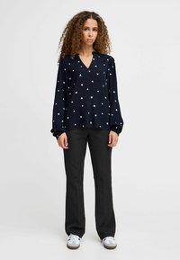 Blouse marine à motif de cœurs blancs, à manches longues et décolleté en V. Associée à un pantalon noir et des baskets blanches. Texture de tissu lisse.