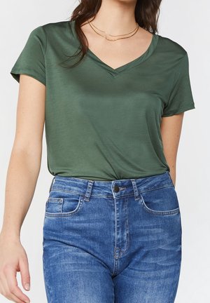Mujer con camiseta verde de manga corta y escote en V metida en unos vaqueros azules de cintura alta con bolsillos delanteros y bolsillo para moneda.
