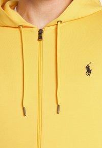 Gul zip-up hoodie i mjukt tyg, med en svart broderad logga, kontrasterande dragsnören och metalliska dragkedjedetaljer.