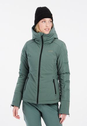 Groene geïsoleerde jas met hoge kraag, ritssluiting aan de voorkant en zijzakken, ontworpen voor warmte. Gecombineerd met een zwarte gebreide beanie.
