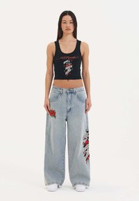 Černý zkrácený top s červeným grafickým designem, v kombinaci s našedlými oversized džíny světle modré barvy, které mají květinové vyšívání a bannerové akcenty. Bílé tenisky.
