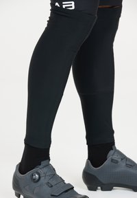 Svarta atletiska benärmar sitter tätt runt underbenen och är tillverkade av ett mjukt, elastiskt material. I kombination med grå cykelskor med snurrstängning.