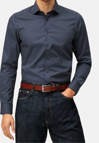 Uomo che indossa una camicia a maniche lunghe blu navy con pois, infilata in jeans di denim scuri, con una cintura di pelle marrone, la mano destra in tasca.