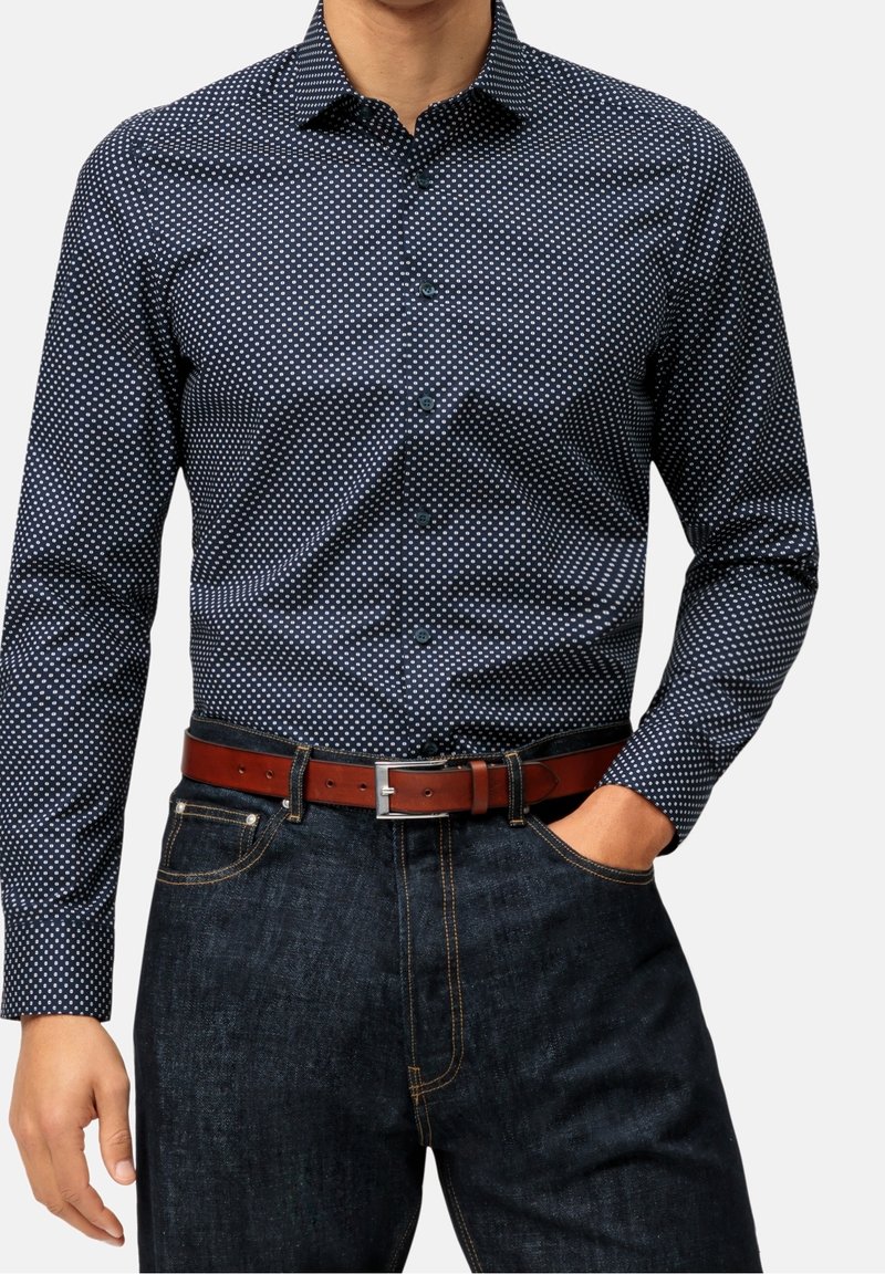 Uomo che indossa una camicia a maniche lunghe blu navy con pois, infilata in jeans di denim scuri, con una cintura di pelle marrone, la mano destra in tasca.
