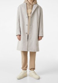 Helles grauer Wollmantel mit spitzem Kragen und drei Knöpfen, über einem beigen Kapuzenpullover und hellbraunen Jogginghosen getragen, kombiniert mit cremefarbenen Sneakers.