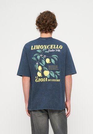 Άνδρας με σγουρά μαλλιά που φοράει μπλε T-shirt με κίτρια λεμόνια, πράσινα φύλλα και ιταλικό κείμενο για το λιμοντσέλο στην πλάτη.