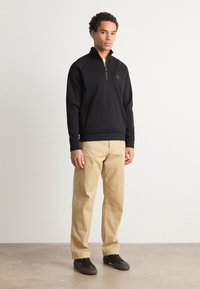Felpa nera a girocollo con zip parziale, corpo texturizzato, polsini a coste e dettaglio del logo, abbinata a pantaloni beige e scarpe scure. Tessuto morbido.