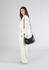 Borsa a tracolla in pelle nera con forma arcuata e accessori dorati, abbinata a una blusa bianca con pizzo e pantaloni a vita alta verde chiaro.