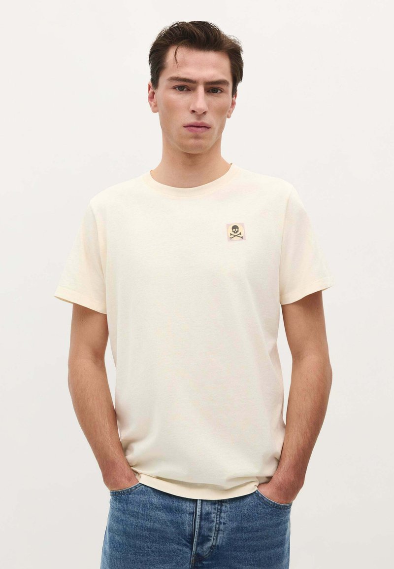 Kortærmet creme t-shirt lavet af bomuld med rund hals og et lille dødningehoved- og korsknogle-mærke på venstre bryst. Casual pasform.