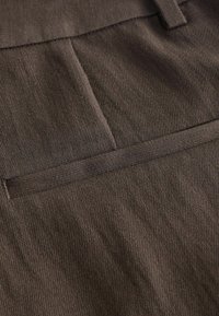 Nahaufnahme von braunem Stoff mit sichtbaren Nähten und einem horizontalen Paspeltaschendetail auf strukturiertem Material.