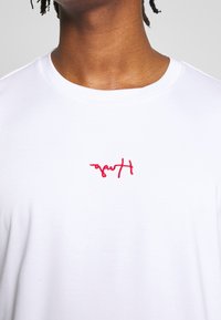 Camiseta de algodón blanca con un logo bordado rojo en la parte frontal. Diseño clásico de cuello redondo, con textura suave y mangas cortas.