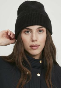 Cappello in maglia a coste nera con vestibilità aderente. Il modello lo indossa con una giacca trapuntata scura in evidenza con dettagli in metallo dorato.