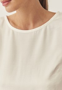 Blusa beige claro con cuello redondo, textura suave y tejido blando que cae con gracia. Carece de patrones o adornos, enfatizando la simplicidad.