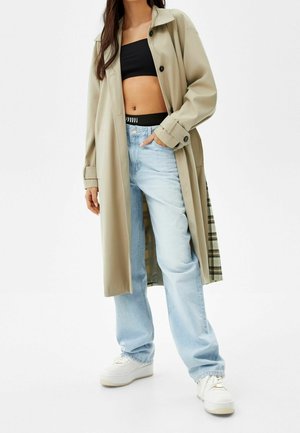 Mujer con gabardina beige, top corto negro, vaqueros azul claro y zapatillas blancas, de pie con una mano en el bolsillo.