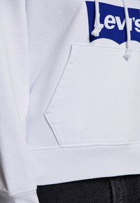 Vit sweatshirt med känguruficka, med en blå Levi's-logotyp tvärs över bröstet. Materialet är en mjuk bomullsblandning med ribbad nederkant.
