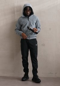 Gray zip-up hoodie with a hood and side pockets, paired with black sweatpants and black sneakers. Holding a black water bottle.