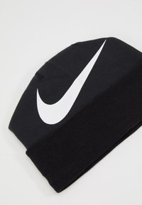 Zwarte gebreide beanie met een dikke boord en een opvallend wit Nike swoosh-logo aan de zijkant. Zachte textuur, veelzijdig ontwerp.