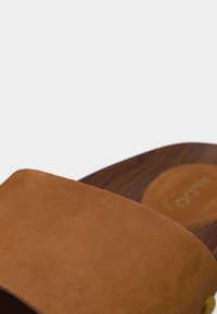 Lanière de sandale en suède marron sur une semelle en bois avec une semelle intérieure rembourrée visible portant la marque "Aldo".