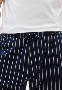 Κοντινή λήψη ατόμου που φοράει ναυτικό μπλε παντελόνι με λευκές κάθετες ρίγες και δεμένο κορδόνι, σε συνδυασμό με ένα απλό λευκό πουκάμισο.