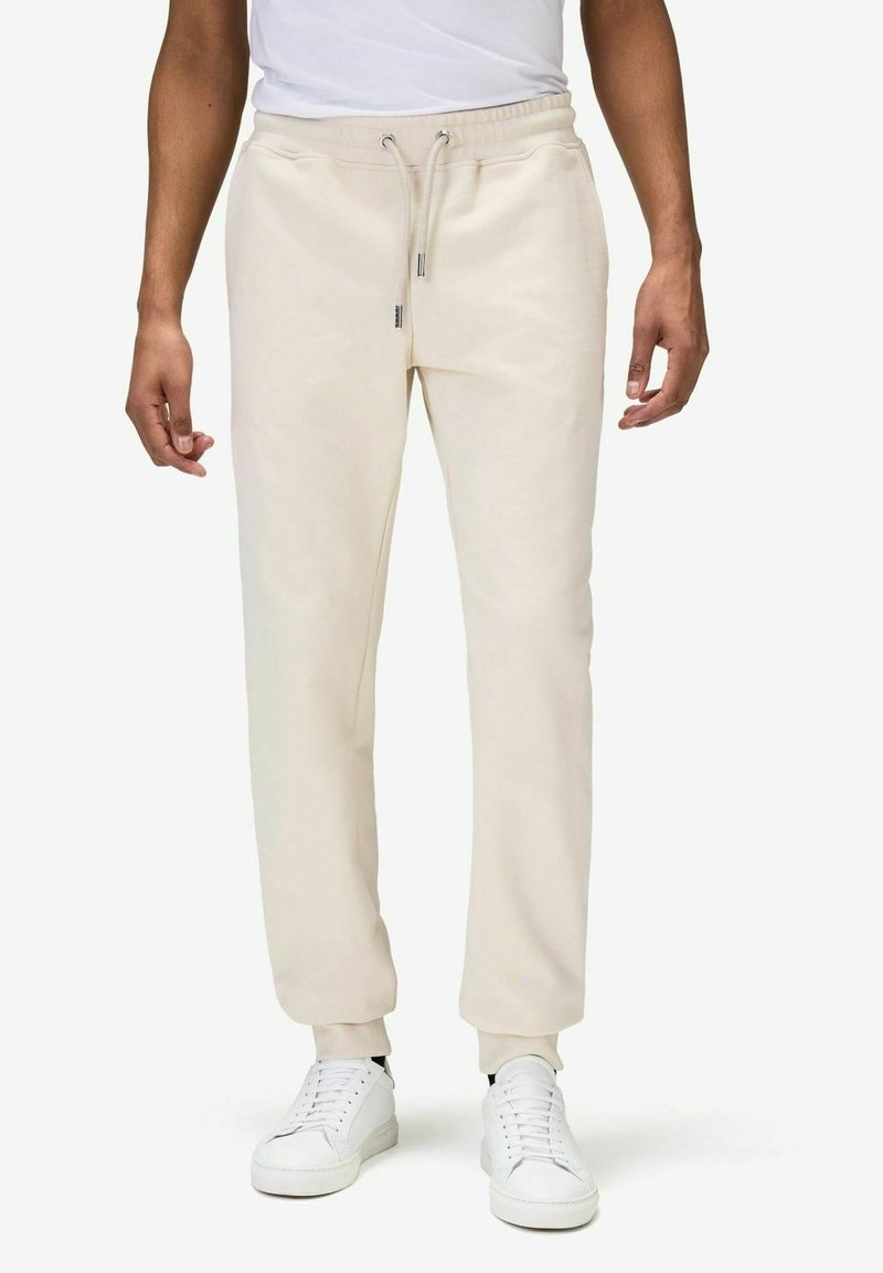 Beige sweatpants med elastisk midjeband, justerbart snöre, sidofickor och avsmalnande nederkant. Bärs med vita sneakers.