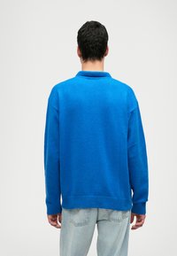 Maglione blu con colletto, realizzato in un tessuto testurizzato. Presenta spalle scese, polsini a coste e orlo dritto, indossato sopra jeans chiari.