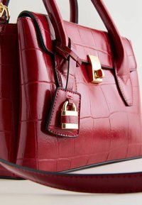 Rote Handtasche aus Krokodilleder mit glänzender Oberfläche, goldfarbener Verschluss, abnehmbares Etikett mit einem Vorhängeschloss-Detail und schwarzer Kante.