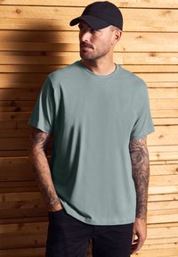 Světle zelené tričko s krátkým rukávem a kulatým výstřihem, s hladkou texturou. Kombinováno s černými kalhotami na pozadí z dřeva.