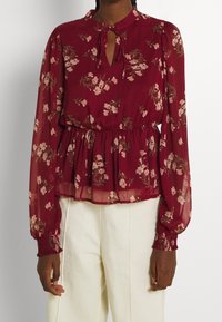 Blouse bordeaux transparente avec motif floral, encolure en trou de serrure, taille froncée et poignets élastiqués. Tissu léger avec un design fluide.