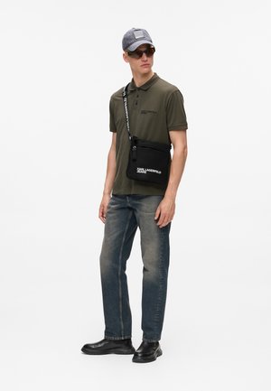 Schwarze Umhängetasche mit Logo, olivgrünes Polohemd, graue Mütze, dunkelblaue Jeans und schwarze Stiefel. Sonnenbrille rundet das Outfit ab.